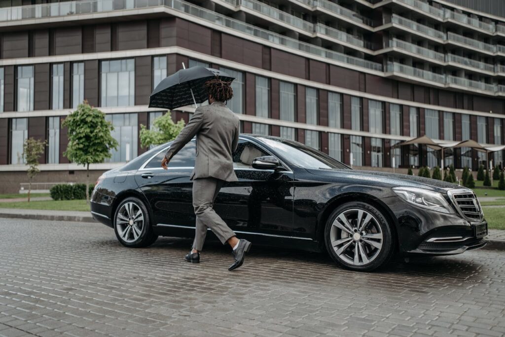 pexels-photo-8425058 Man holding umbrella opens car door on rainy day in front of modern building.
